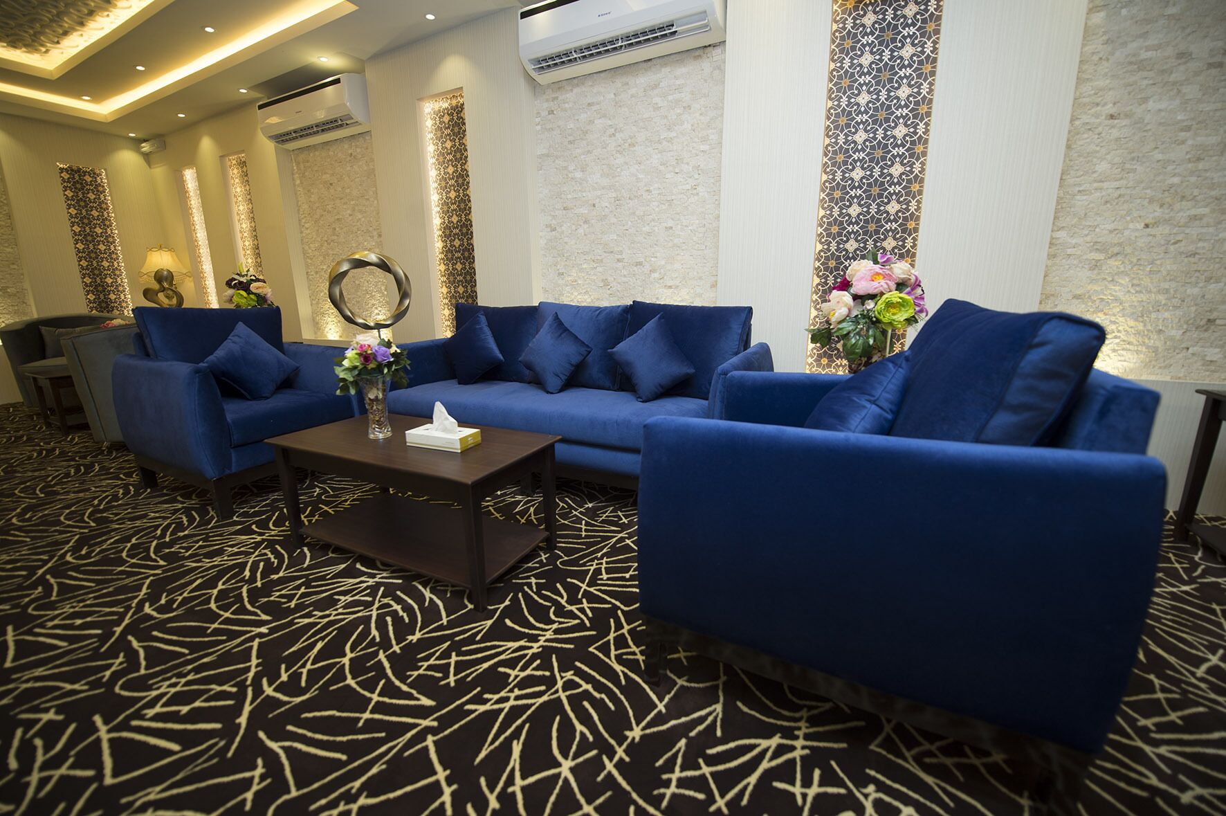 Un moderno espacio de descanso con un sofá azul, una mesa de centro y detalles decorativos como un jarrón con flores y un espejo montado en la pared.