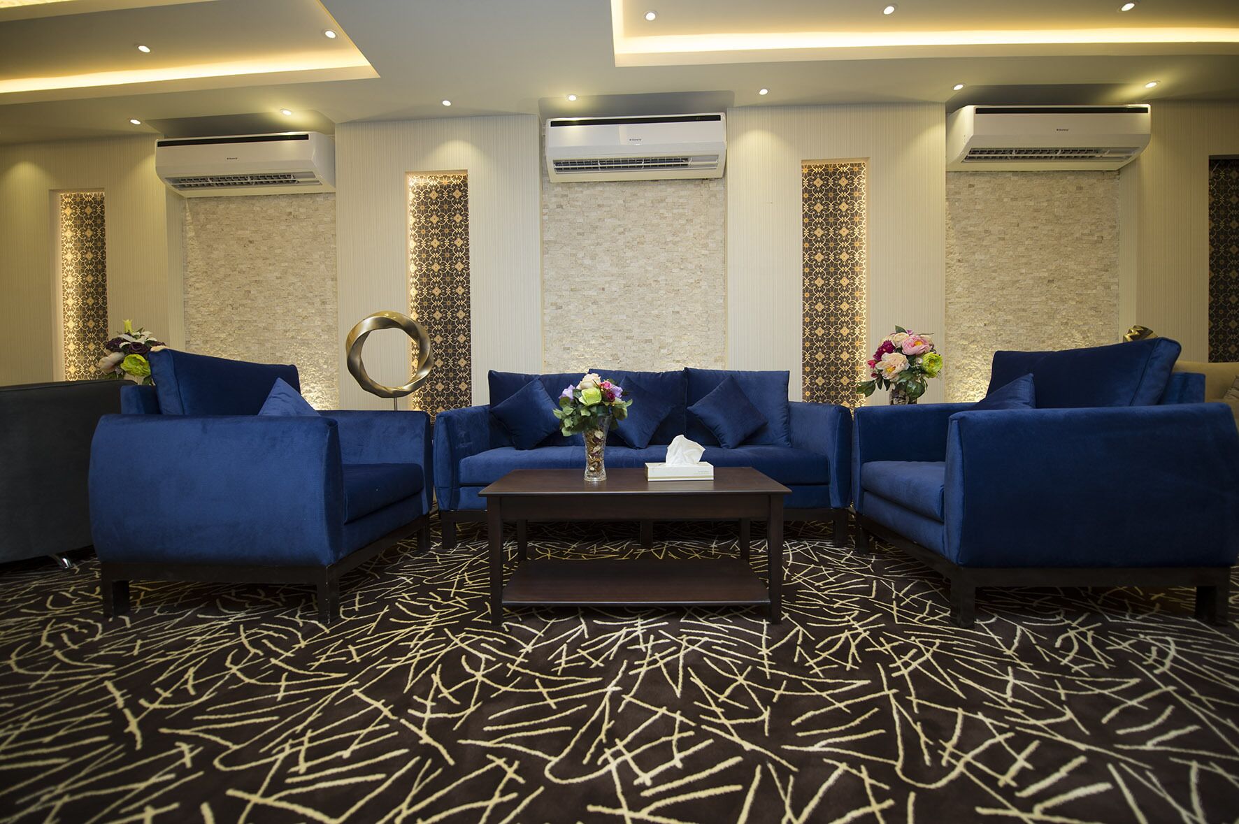 A modern lounge area with blue sofas, a central coffee table, and decorative wall panels.
