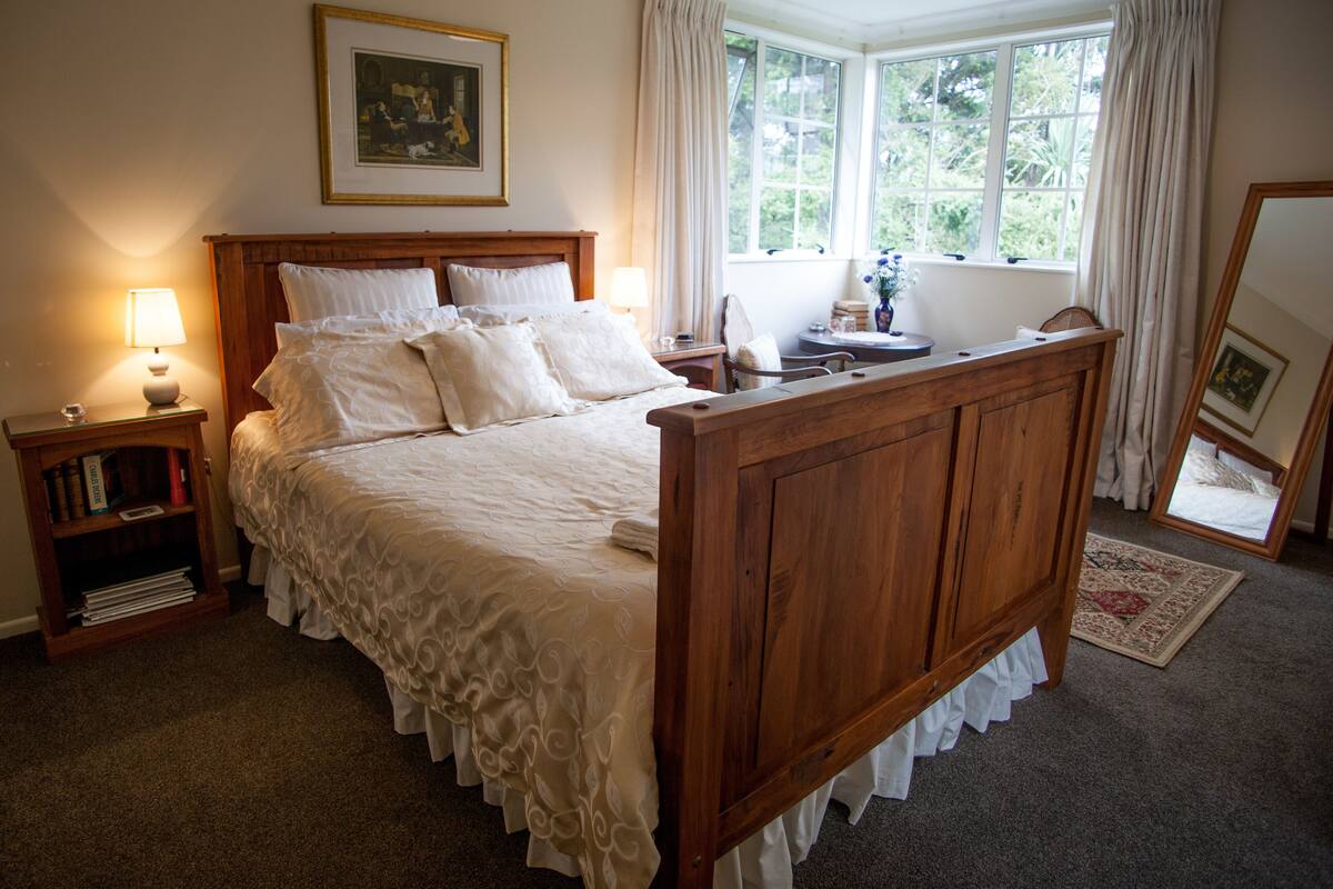 A bedroom with a wooden bed, a bedside table with a lamp, a nightstand with books, a window with curtains, and a framed picture on the wall.