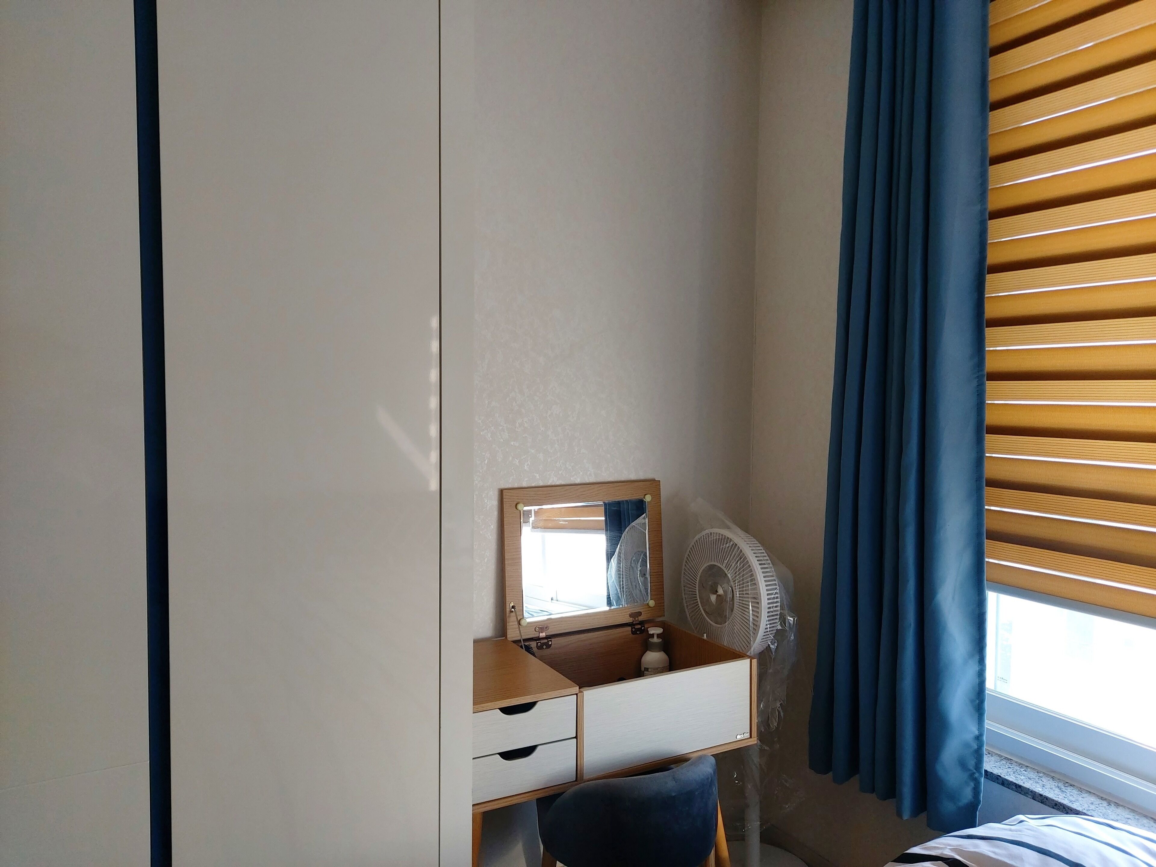 A bedroom with a wooden dresser, a mirror, a fan, and a window with blinds.