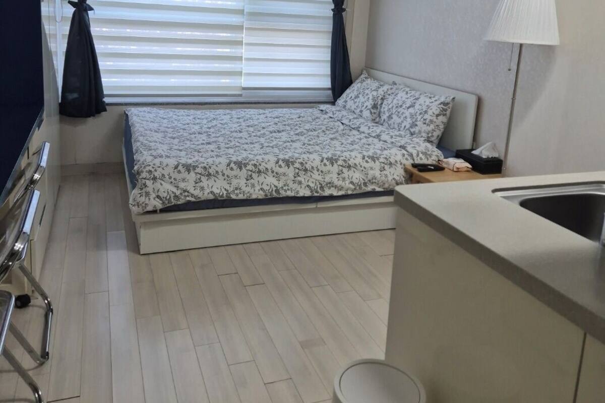 Una habitación pequeña con cama, mesita de noche, lámpara, lavamanos y ventana con persianas.