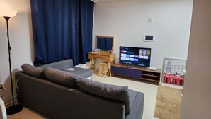 Una sala de estar moderna con un sofá gris, un escritorio de madera, un televisor mostrando servicios de transmisión y un gran ventanal con cortinas.