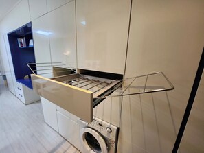 Una cocina compacta con lavadora integrada, un tendedero y una pequeña estantería.