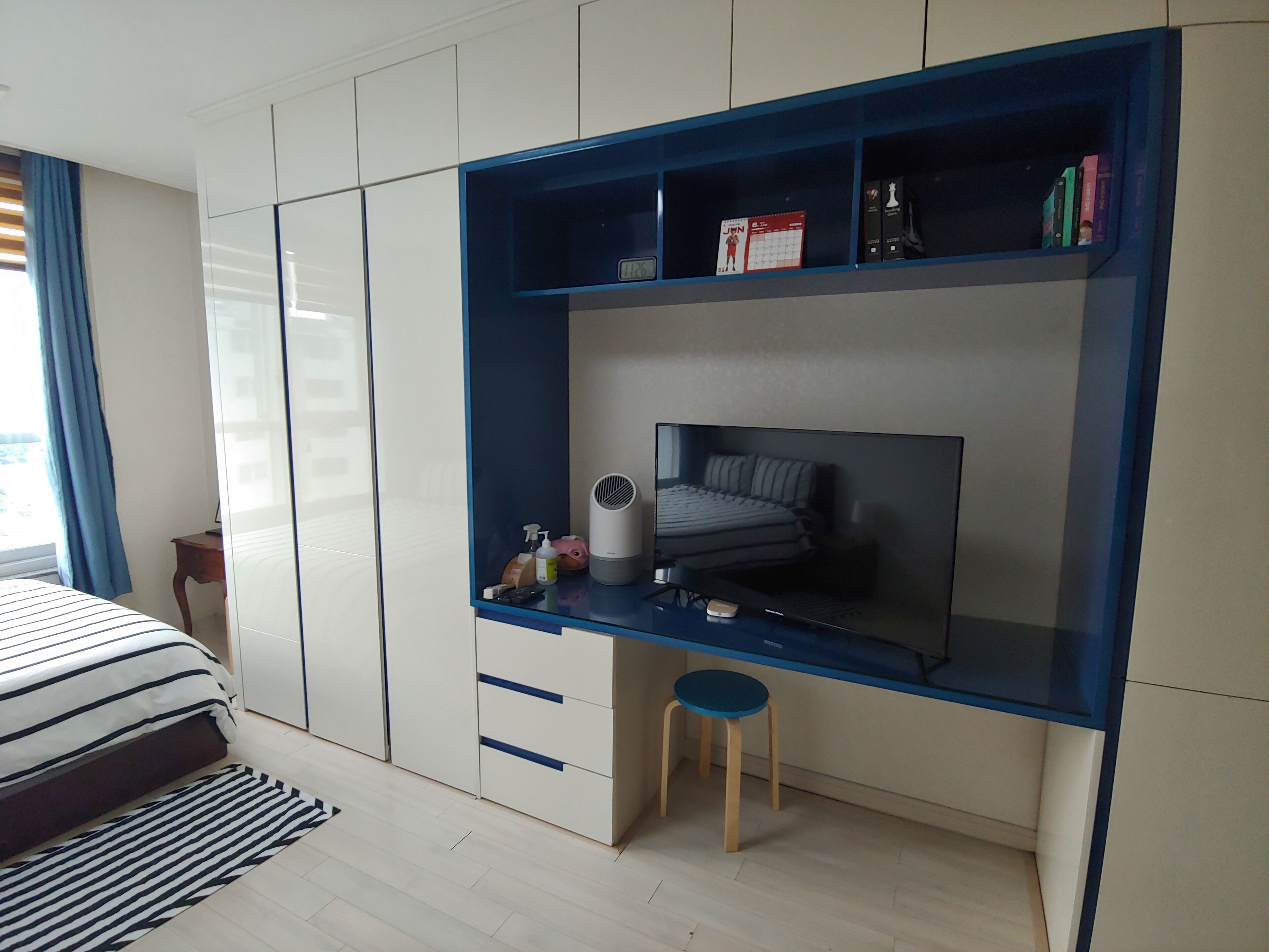 Un dormitorio con una cama, un escritorio con silla, un taburete pequeño y una estantería con libros y un calendario.