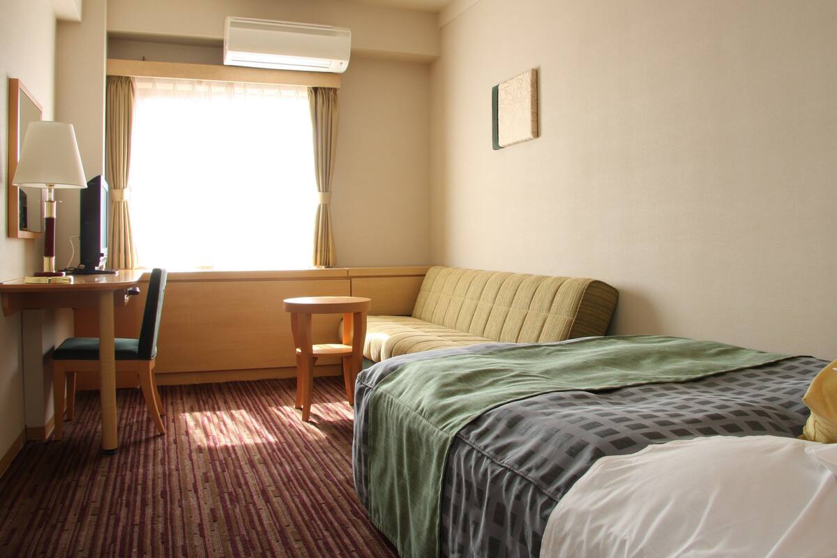 Ein Hotelzimmer mit einem Bett, einem Schreibtisch mit Stuhl, einem kleinen Tisch und einem Fenster mit Vorhängen.