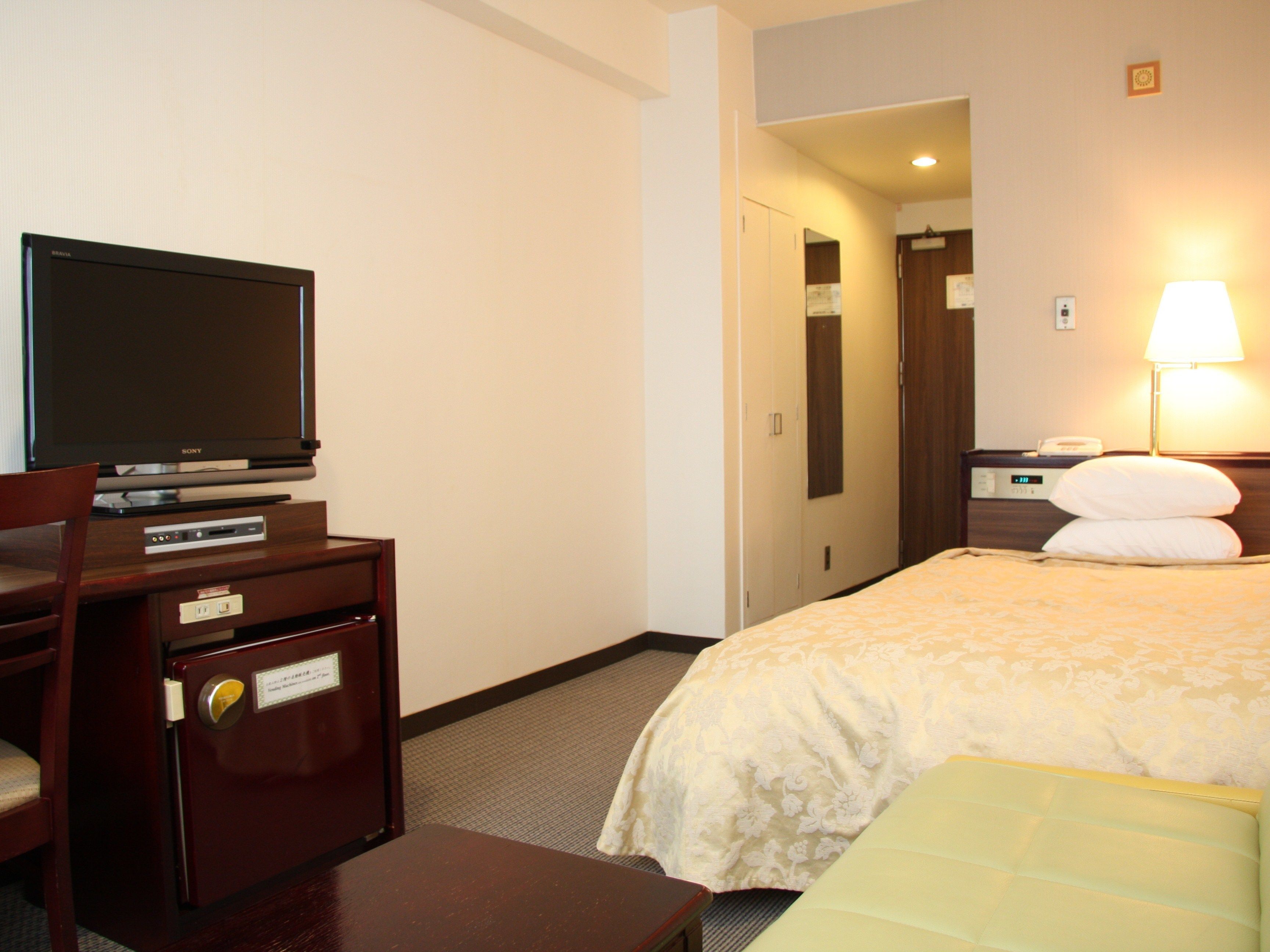 A hotel room with a bed, a desk with a television, a chair, a bedside table with a lamp, and a nightstand.