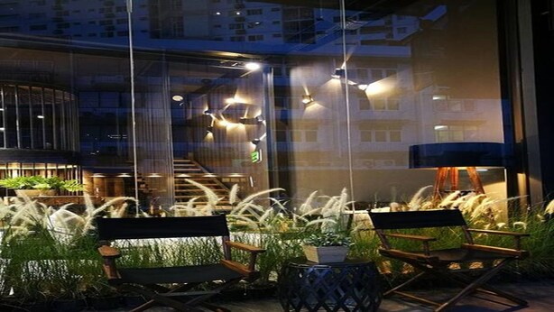 An outdoor seating area with modern chairs and a small table, surrounded by tall grasses and plants, with a building and staircase in the background. - Sook Station (Bangkok)