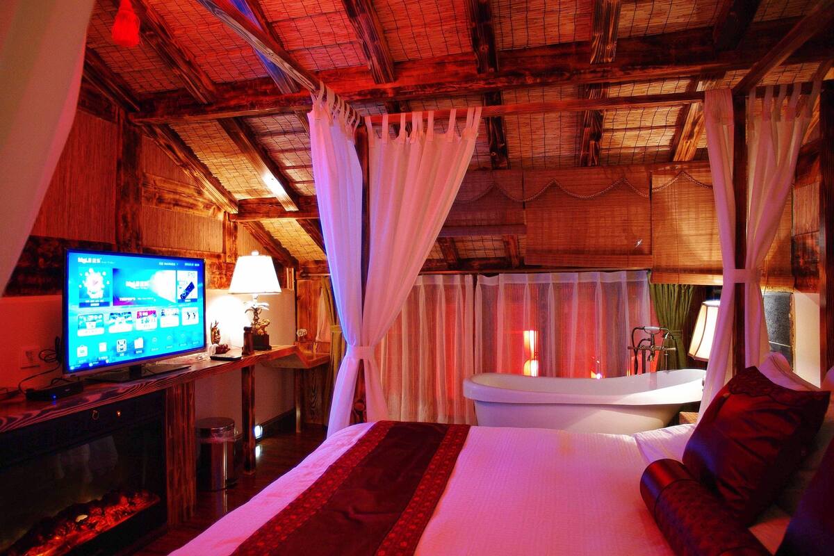 A room with a bed, a television, a bathtub, and a window with curtains.