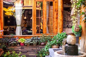 A wooden house with a large window and a garden area with various potted plants and a stone fountain.