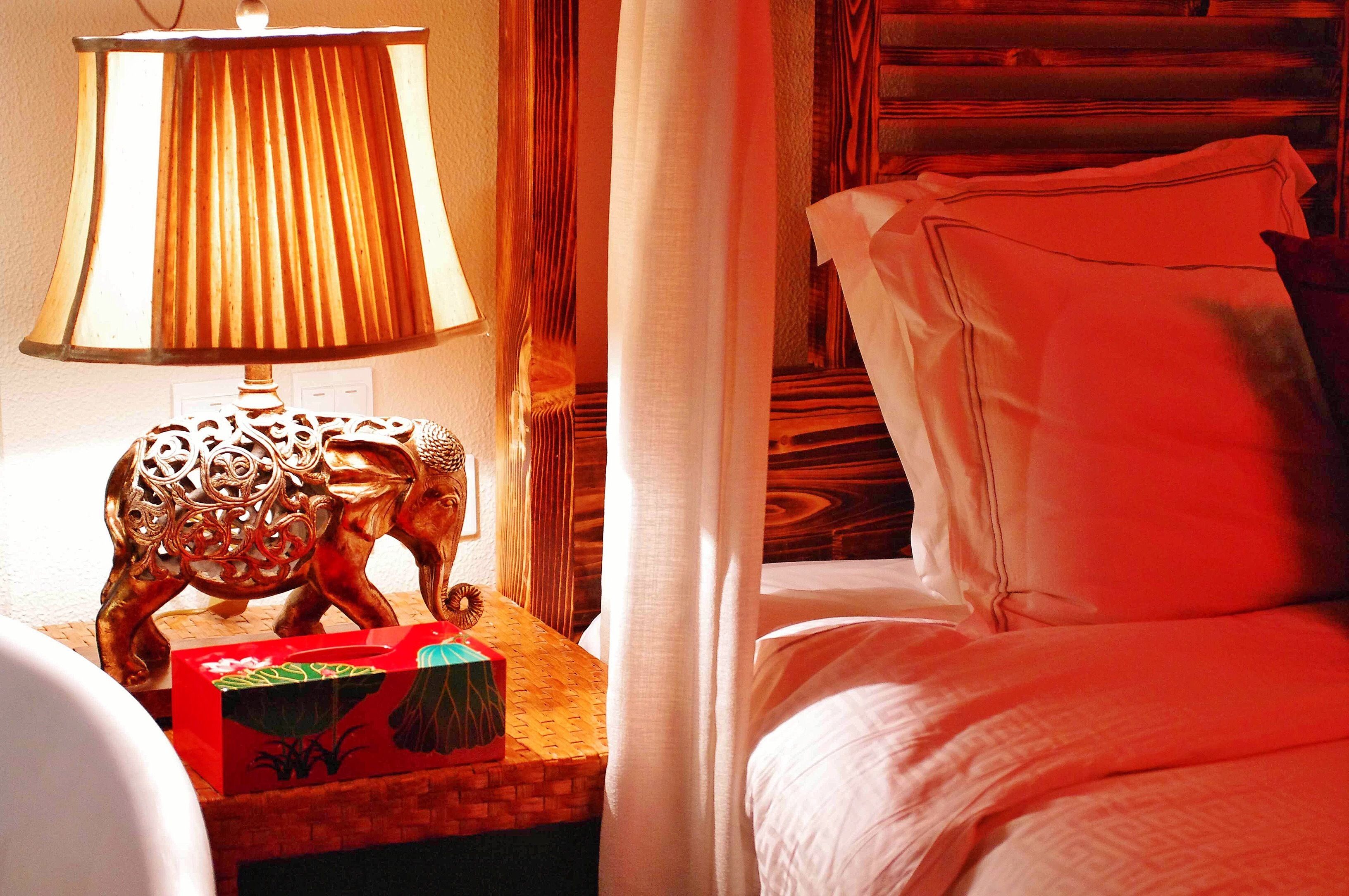 Un lit bien fait, avec du linge de lit blanc et des oreillers, une lampe décorative surmontée d’un pied en bois, et une petite table sur laquelle se trouvent une boîte et un objet décoratif.