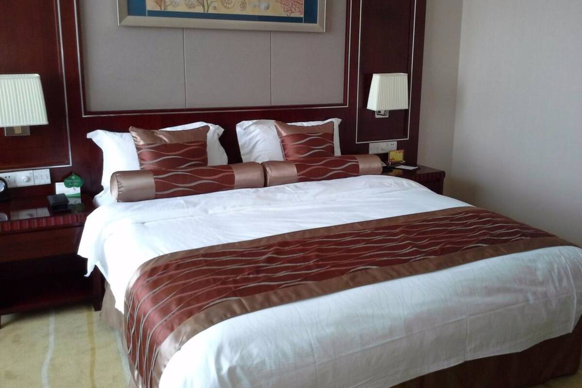 Une chambre d’hôtel avec un grand lit, deux tables de chevet, une petite table d’appoint, une lampe fixée au mur et une œuvre d’art encadrée au-dessus du lit.