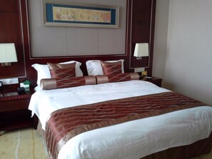 A hotel room with a large bed, two bedside tables, a nightstand, a wall-mounted lamp, and a framed artwork above the bed. - Qingdao Huaxi Hotel (Qingdao)