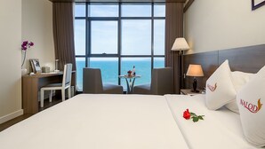 Habitación de hotel con una cama grande, un escritorio, una silla, una mesita con una copa de vino y vista al mar.
