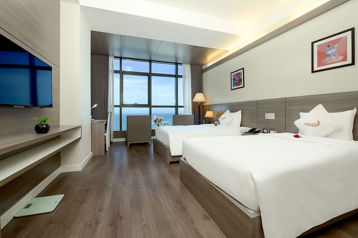 A hotel room with two beds, a desk, a TV, and a large window.