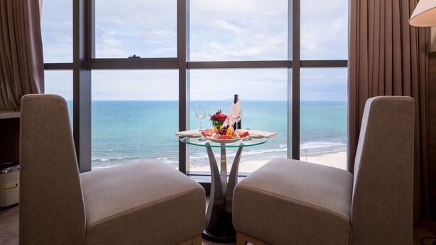 Una habitación con un amplio ventanal con vista a la playa, una mesa de vidrio con una botella, copas de vino y fruta, y dos sillones color beige.