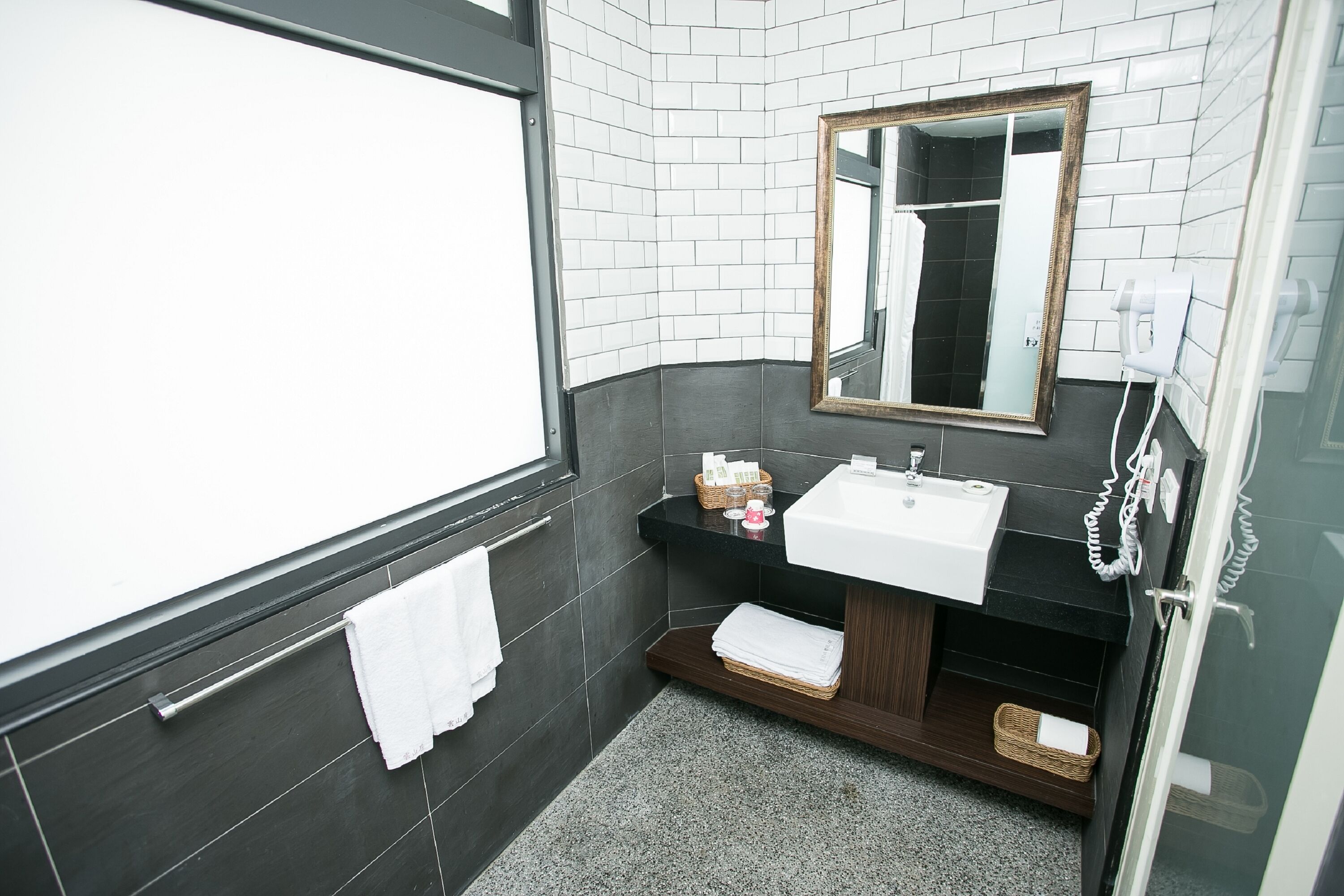 A modern bathroom with a white sink, dark tiled walls, a wooden vanity, a mirror, and a glass shower enclosure.