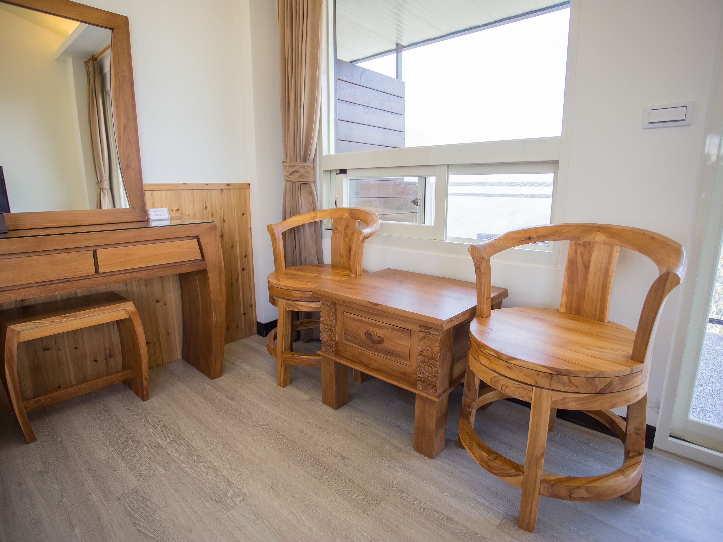 Ein Holztisch mit Schubladen, ein Stuhl und ein runder Holzschemel in einem Zimmer mit Fenster und Spiegel.