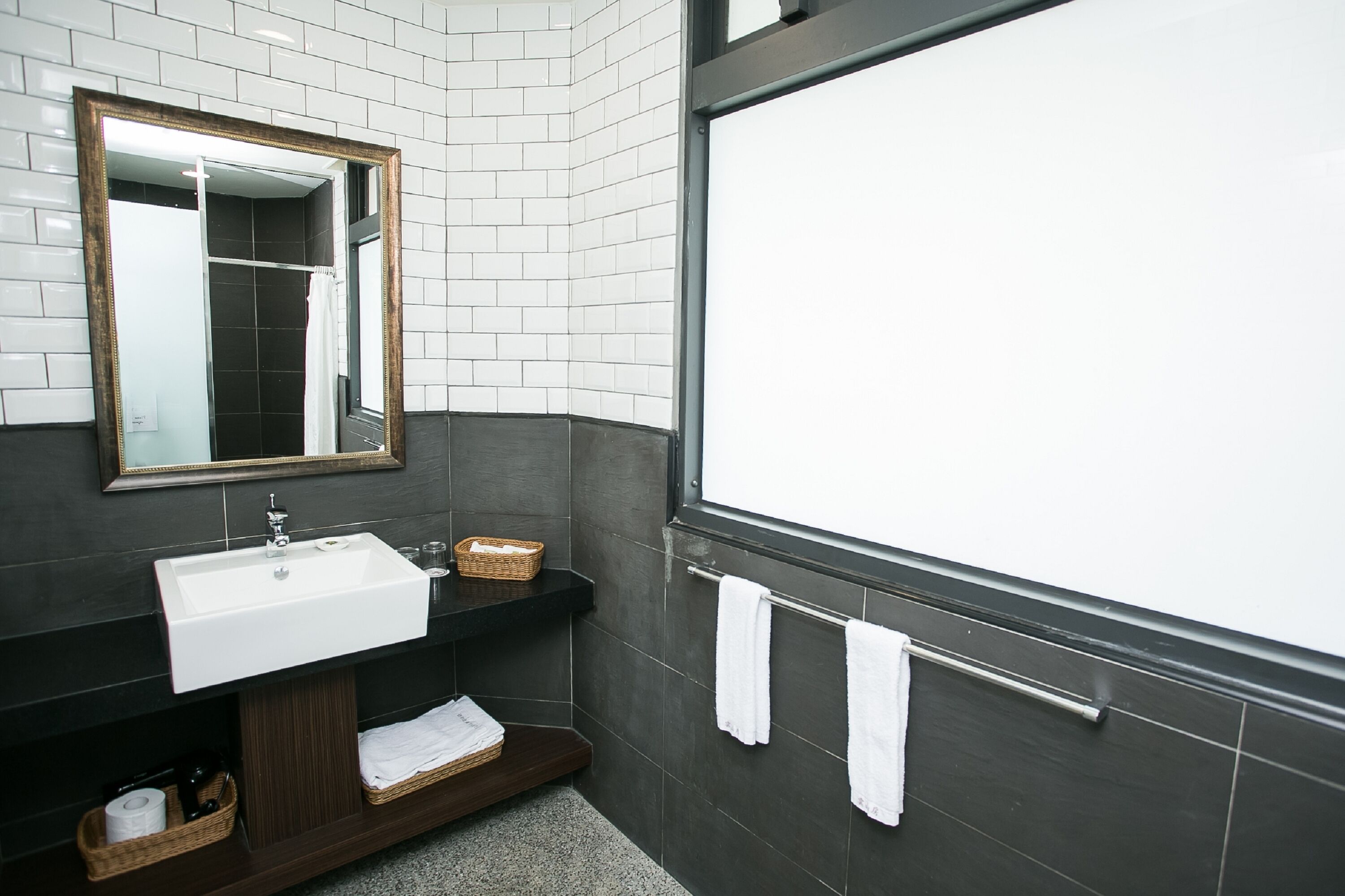 Un baño moderno con un lavabo blanco, baldosas oscuras, un espejo grande y un perchero para toallas.