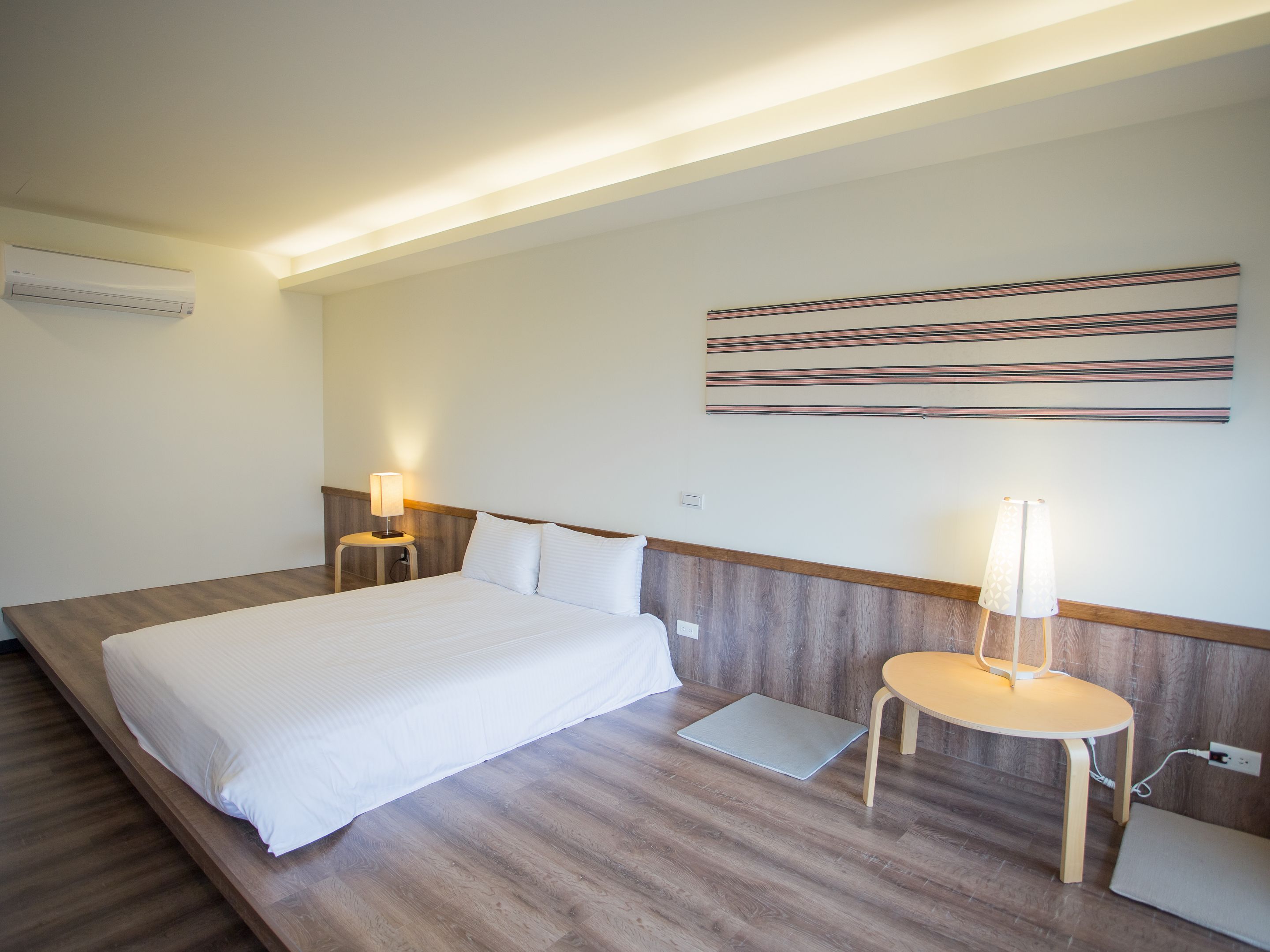 A hotel room with a large bed, two bedside tables with lamps, a laptop on the floor, and a wall-mounted air conditioner.