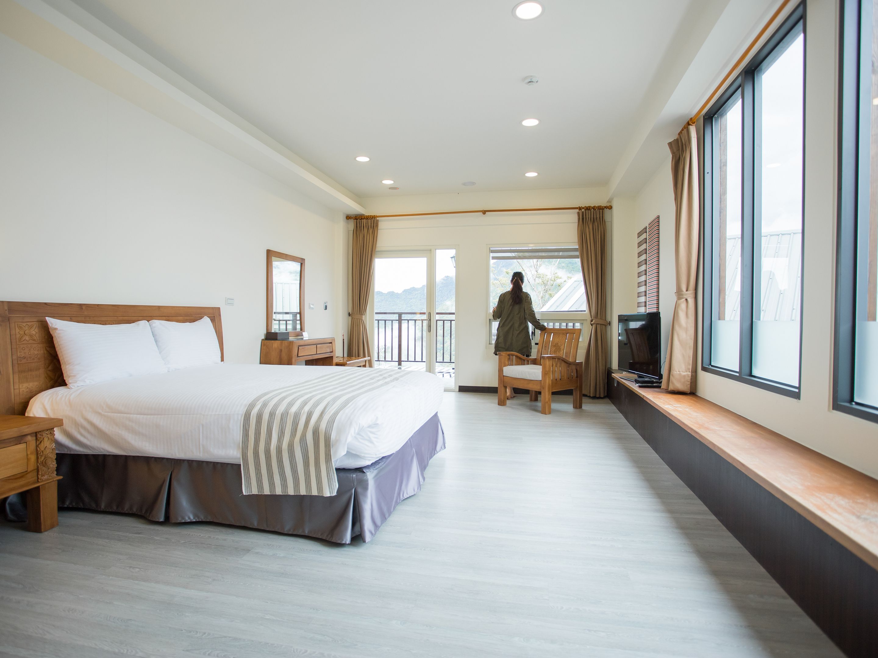 A hotel room with a large bed, a wooden nightstand, a desk with a chair, a TV, and a balcony with a view of the mountains.