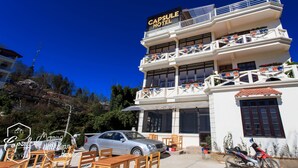 Un hotel de varios pisos con balcones, un coche estacionado adelante y mobiliario exterior.