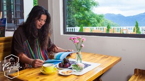 Una persona leyendo en una mesa de madera, con una taza amarilla, un plato de comida y un florero con flores.