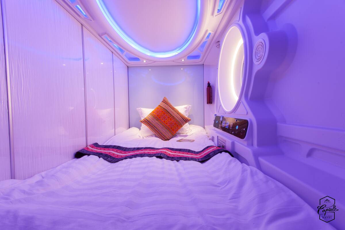 A modern, well-lit bedroom with a neatly made bed, a bedside table with a clock, and a circular light fixture above the bed.
