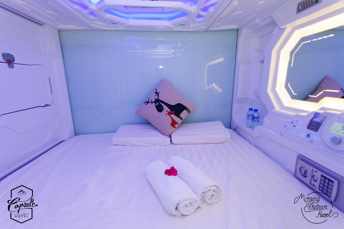 A modern hotel room with a neatly made bed, a pillow with a reindeer design, and a control panel on the side.