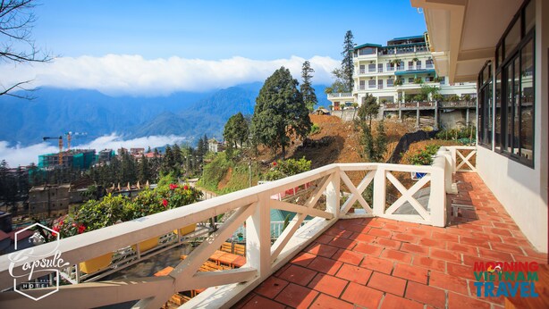 Un balcón de hotel con suelo de tejas rojas, baranda blanca y vistas a montañas y nubes.