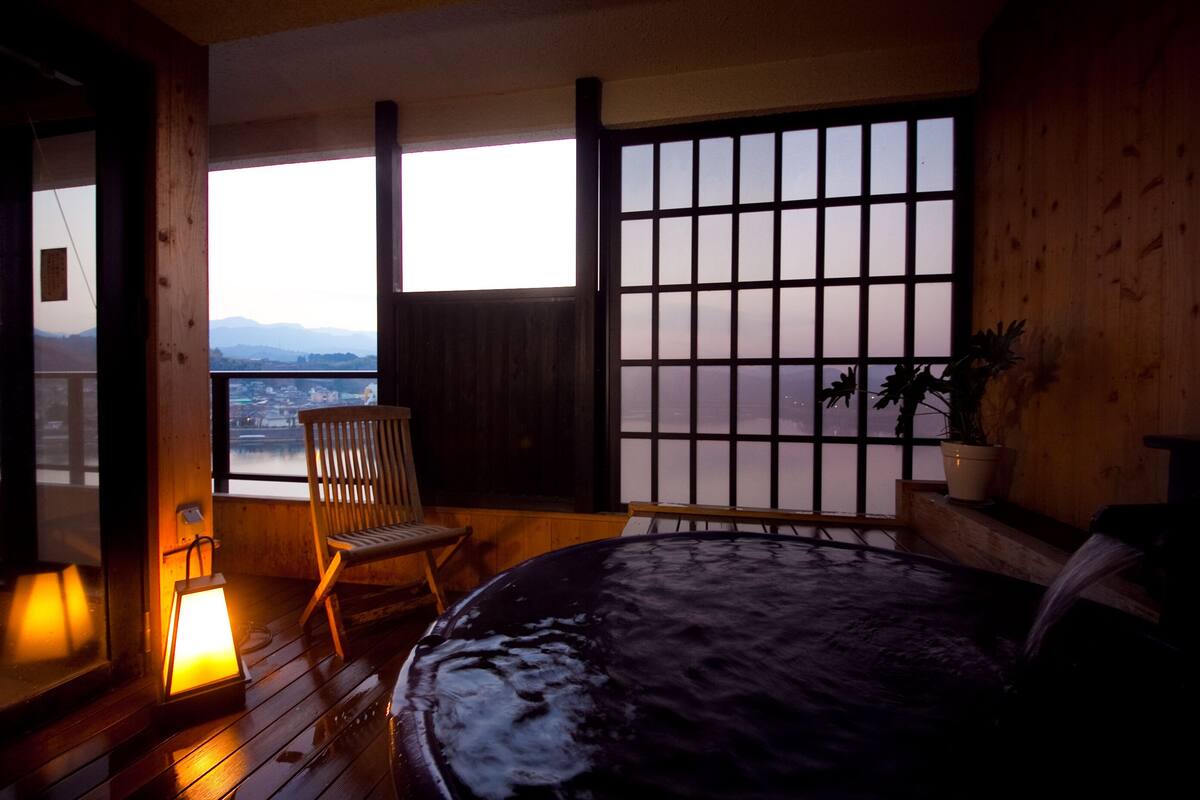 A room with a bed, a wooden chair, a lantern, and a view of mountains through a window.