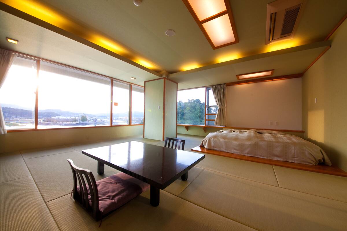 A room with tatami flooring, a low table, chairs, a bed, and large windows offering a view of the outdoors.