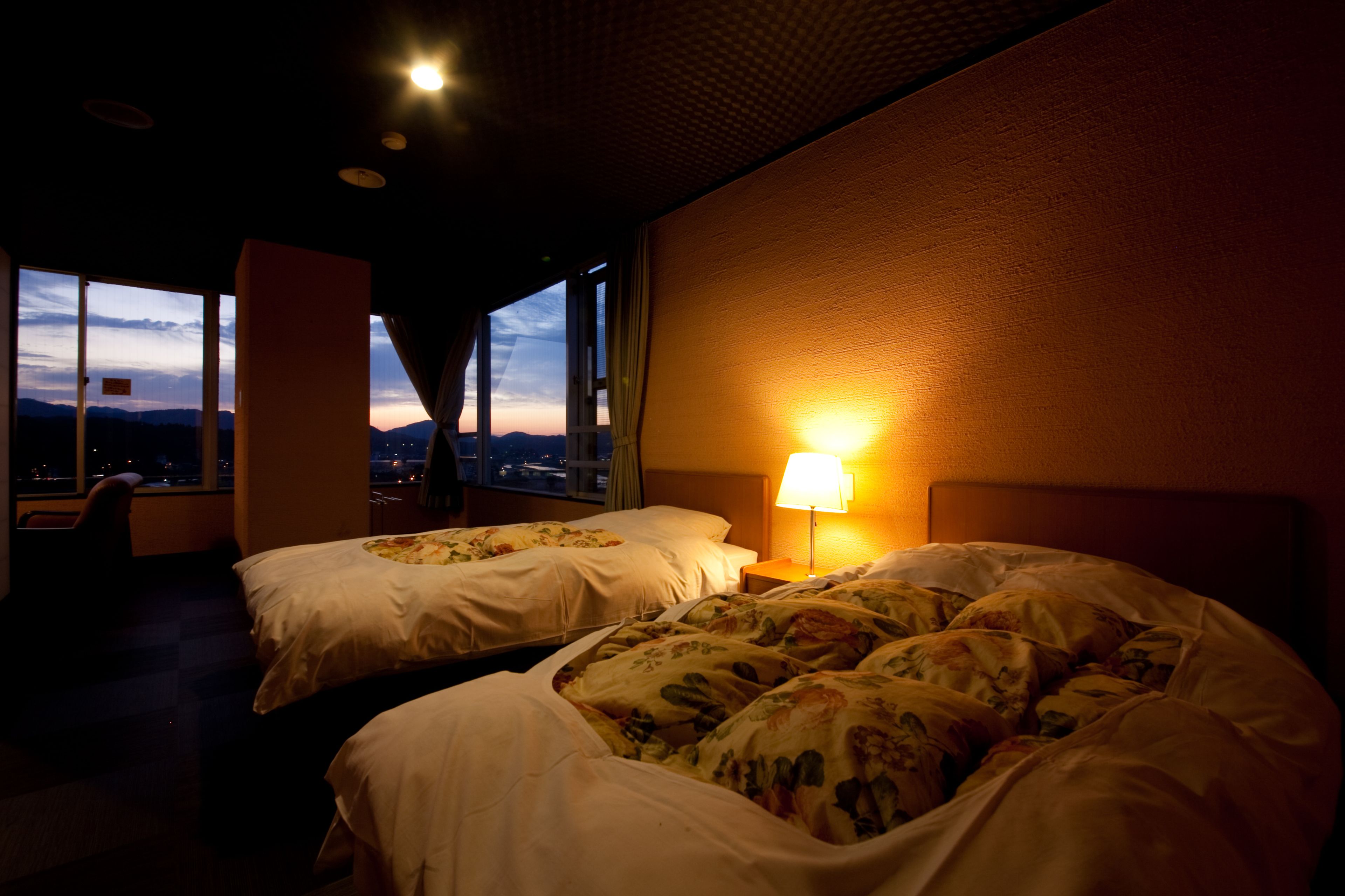 A hotel room with two beds, a nightstand with a lamp, and a view of the city at dusk.
