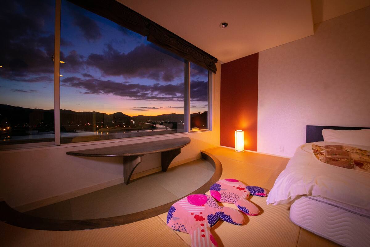 A modern hotel room with a large window, a bedside table, a bed, and a unique patterned rug.