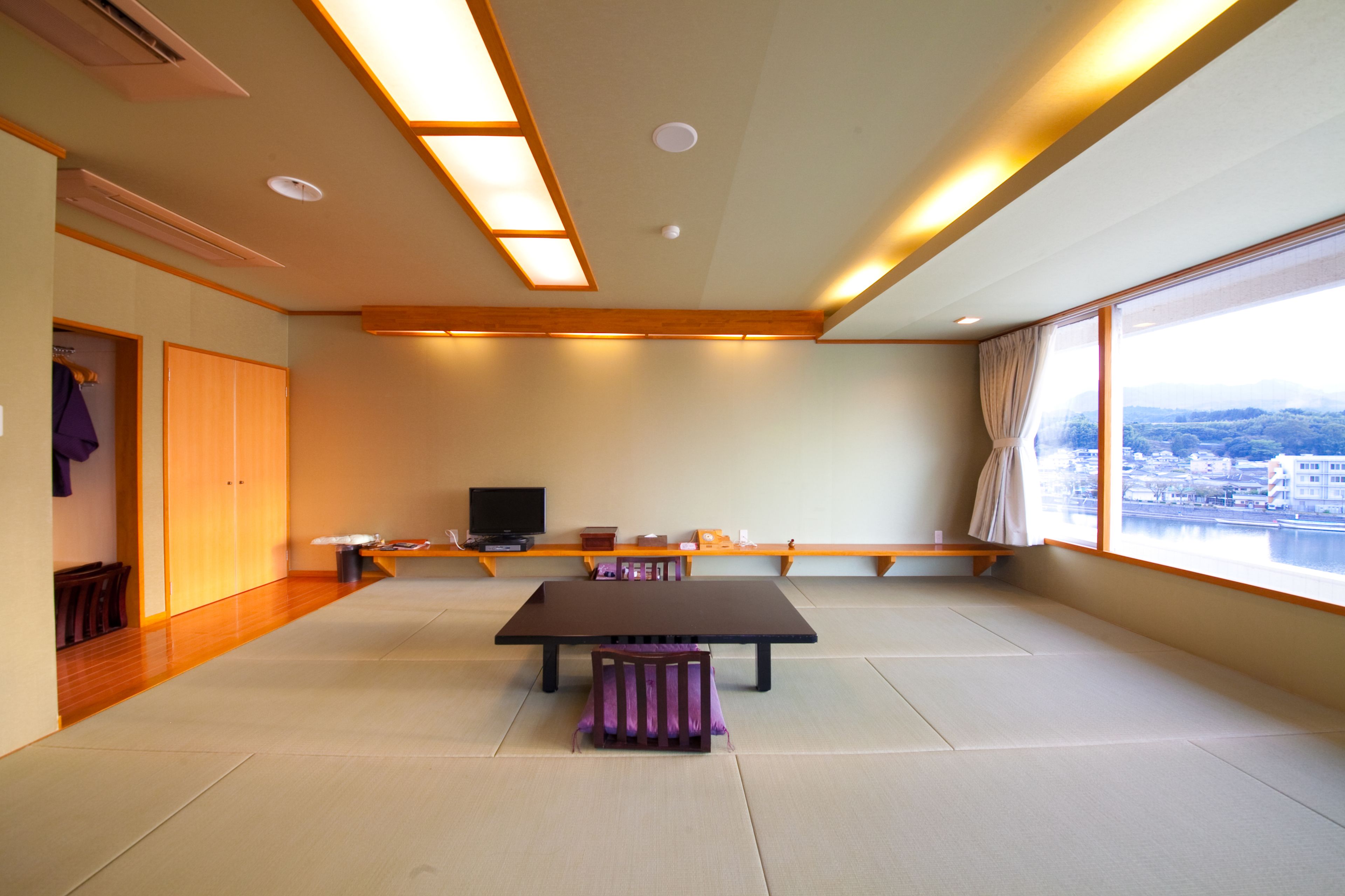 Amplia habitación con suelo de tatami, una mesita baja, televisor y un ventanal con vistas a la ciudad.