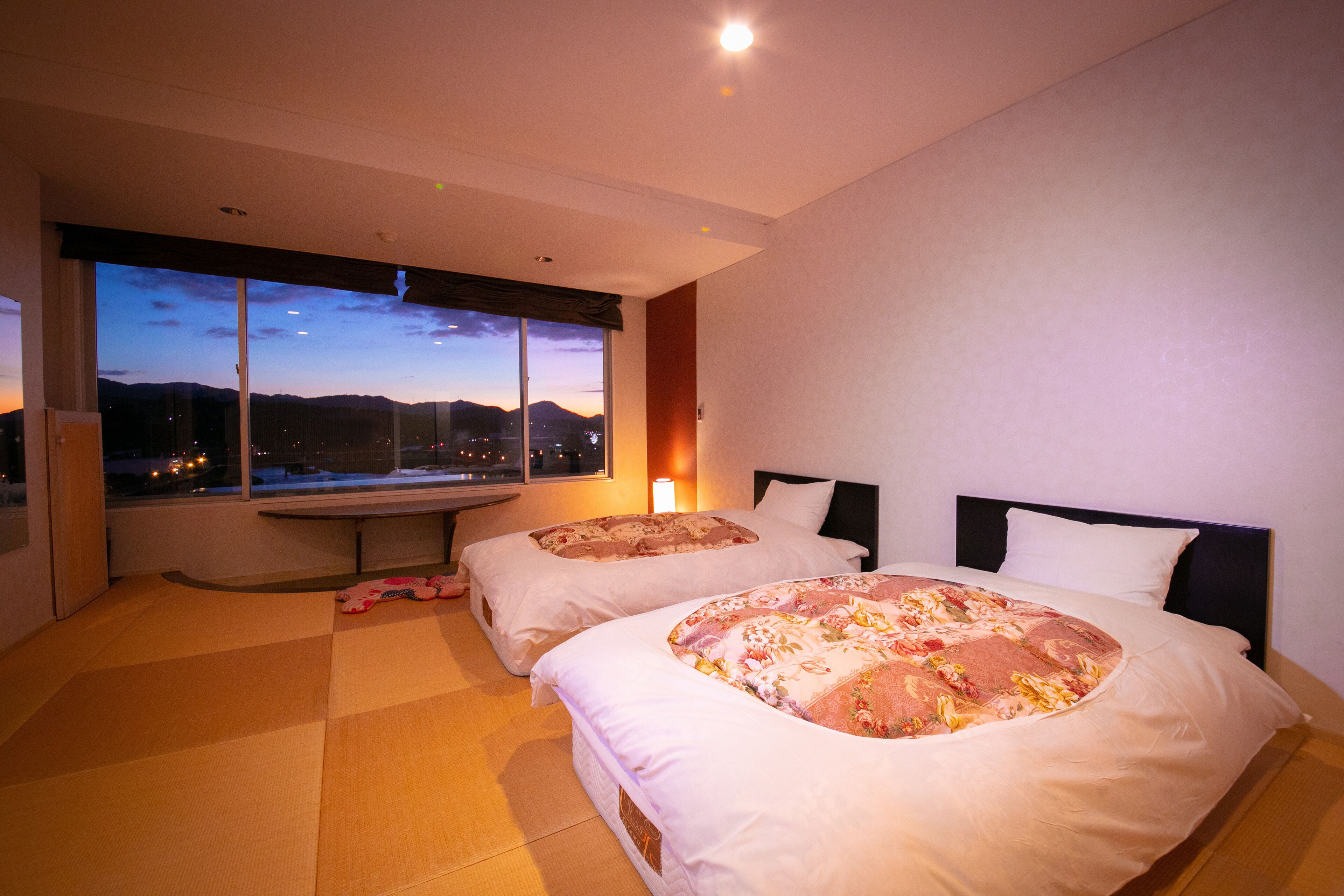 Habitación de hotel con dos camas, un ventanal con vistas a la ciudad al anochecer y un banco junto a la ventana.