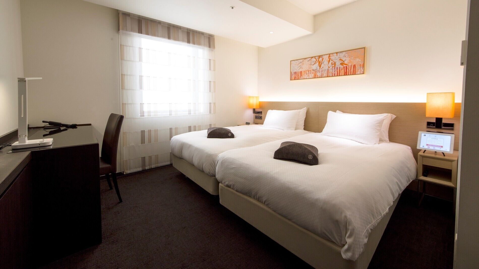 A hotel room with two beds, a desk, a chair, a window with blinds, a painting on the wall, and two bedside lamps.