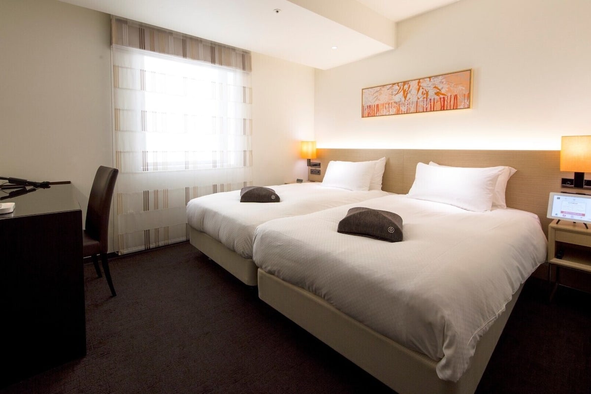 A hotel room with two beds, a desk, a chair, a window with blinds, a painting on the wall, and two bedside lamps.