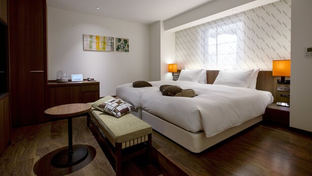 Quarto de hotel moderno com uma cama grande, abajures, uma mesinha e um banco.