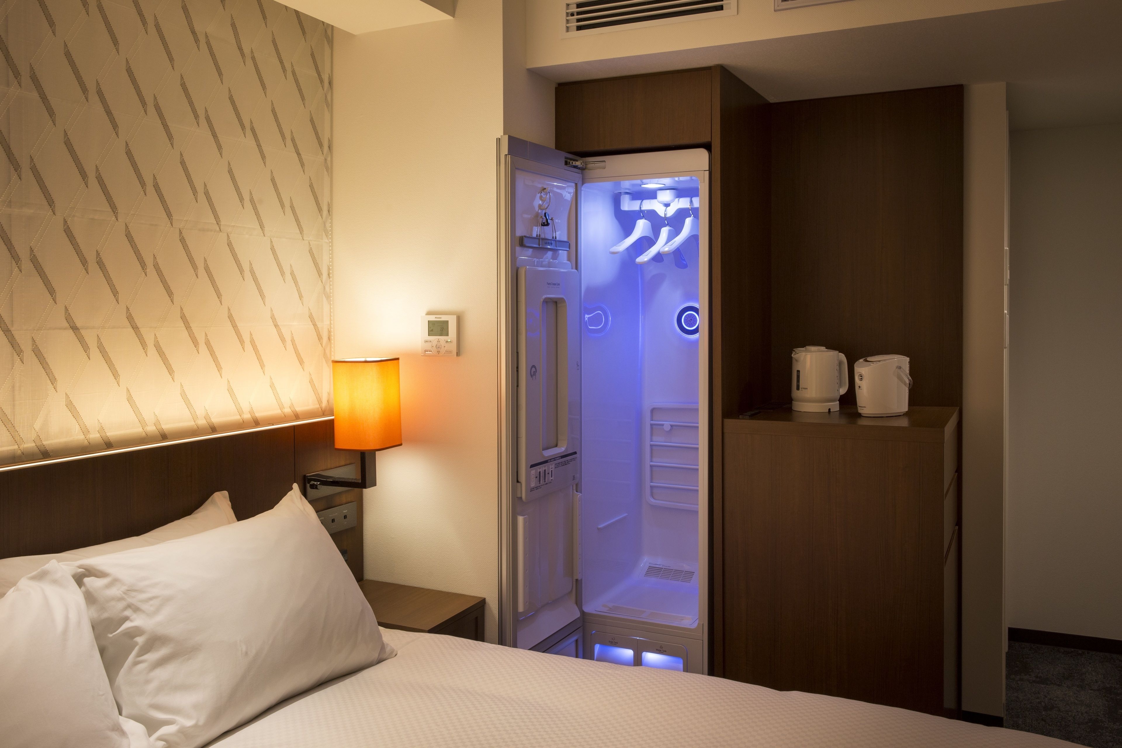 A hotel room with a bed, a nightstand with a lamp, a minibar, and a built-in refrigerator.