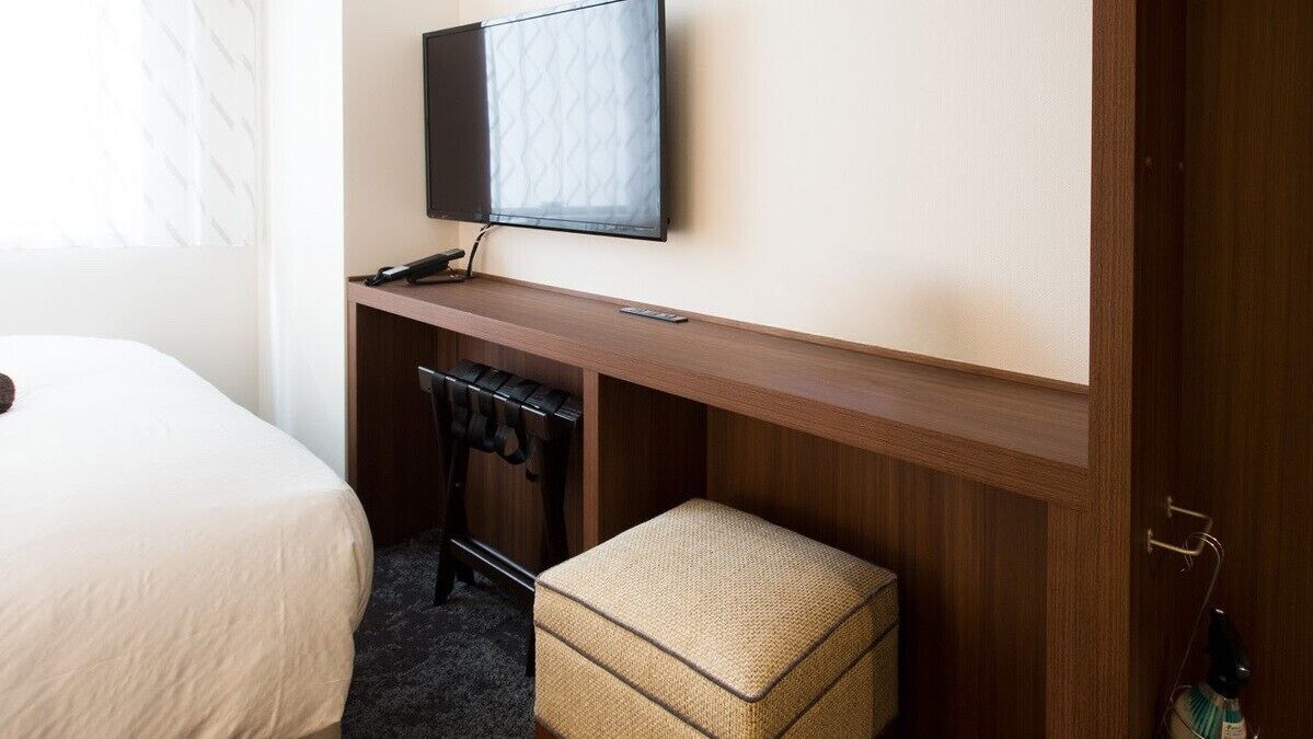 A hotel room with a bed, a desk with a television, a chair, and a small ottoman.