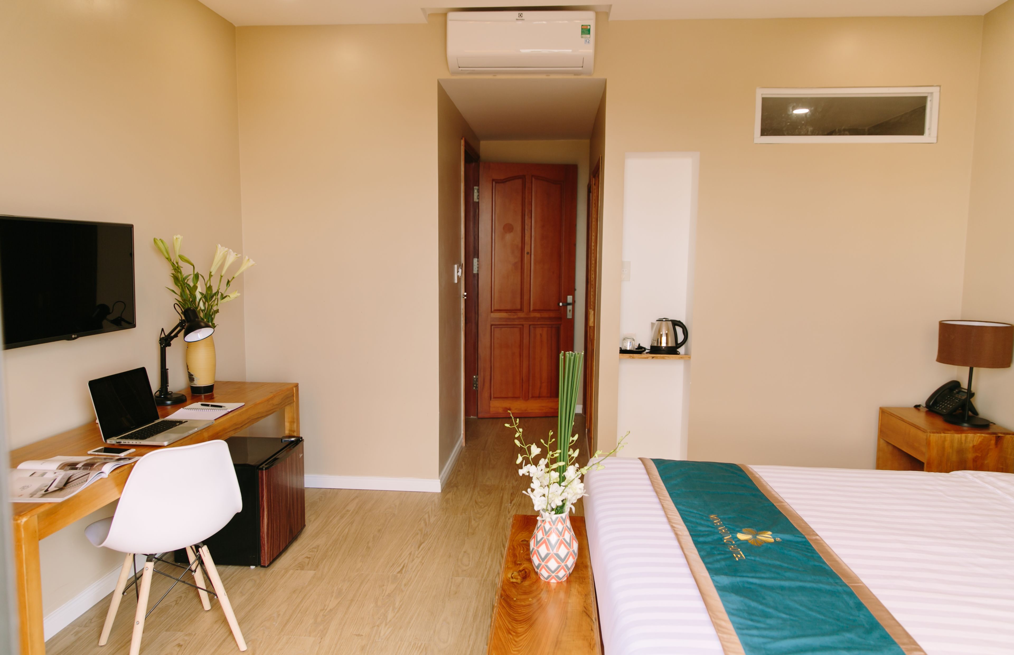 Habitación de hotel con cama, escritorio, silla, televisión y una pequeña mesa con lámpara y flores.