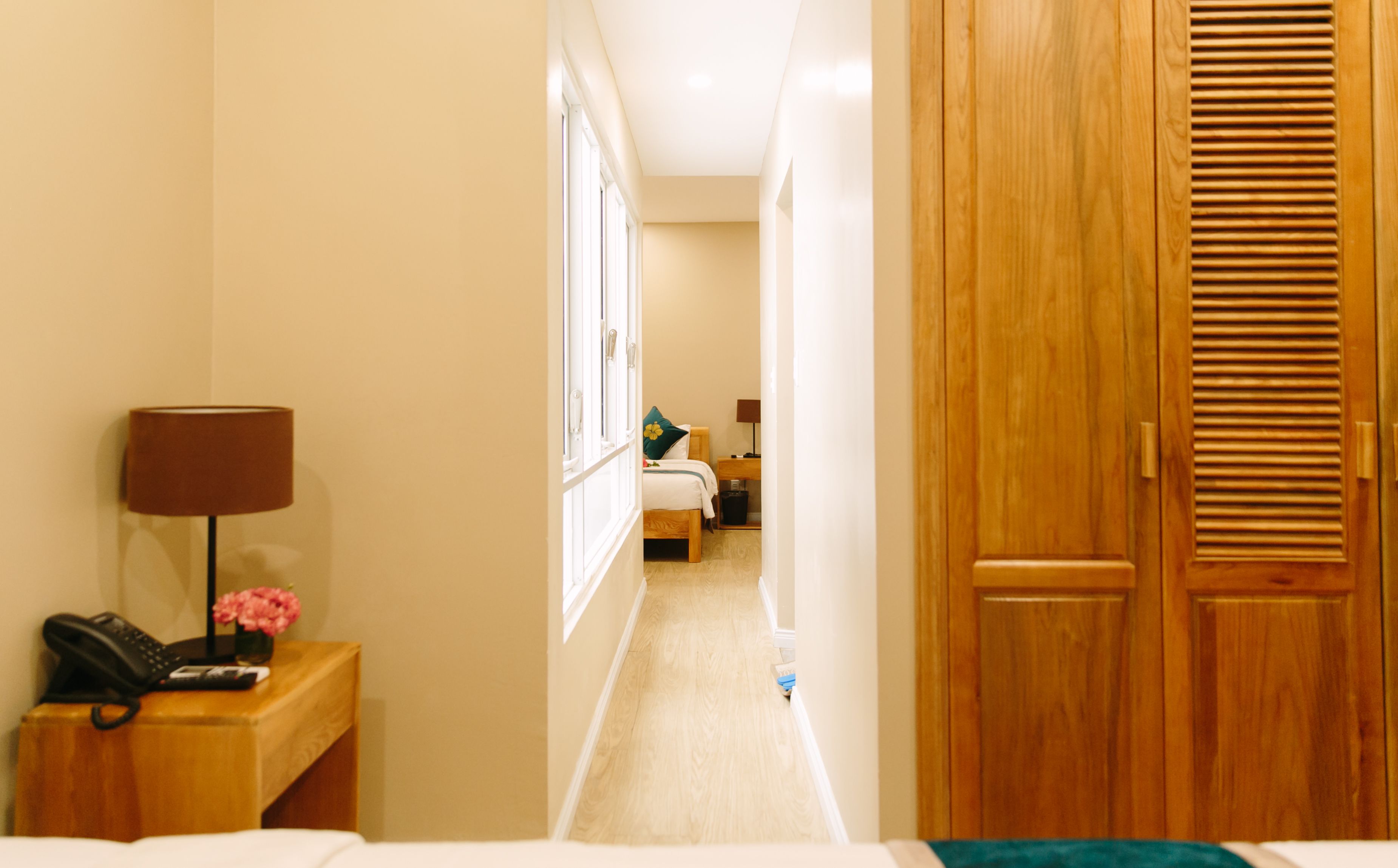 Un estrecho pasillo de hotel, con una cama a la izquierda, una puerta de madera a la derecha y una mesita de noche con una lámpara y teléfono.