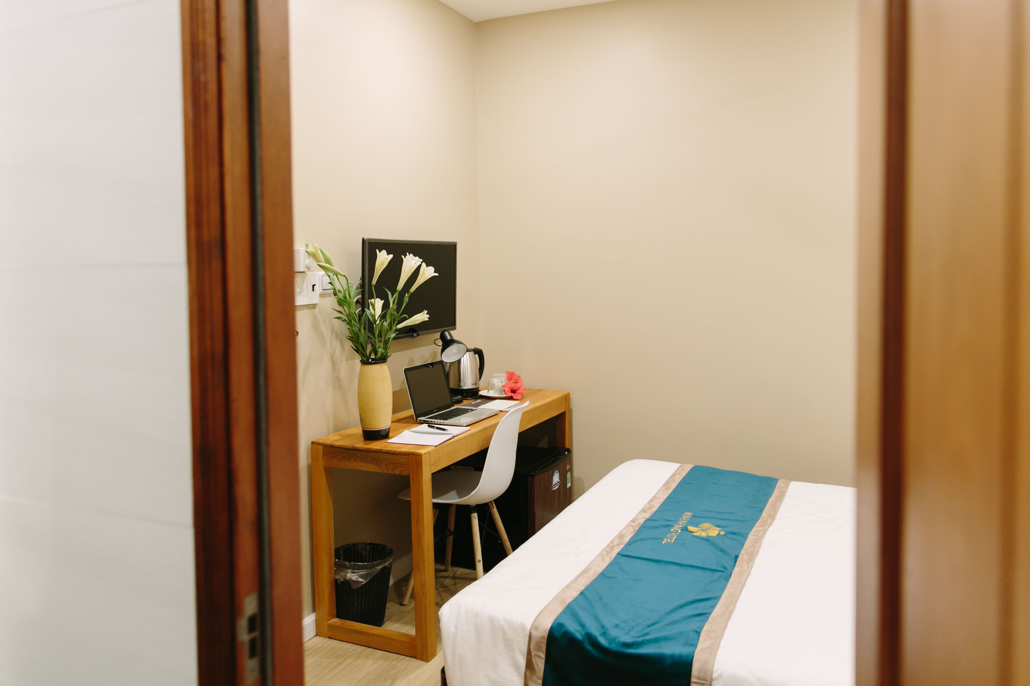A hotel room with a bed, a desk with a laptop, a chair, a potted plant, and a small trash bin.
