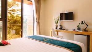 A hotel room with a bed, a desk with a computer, a chair, a TV, and a window with a view of trees. - Minh Nhung Hotel (1 Bao Loc)