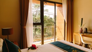 A hotel room with a bed, a desk, a chair, a lamp, and a view of trees through the window. - Minh Nhung Hotel (1 Bao Loc)