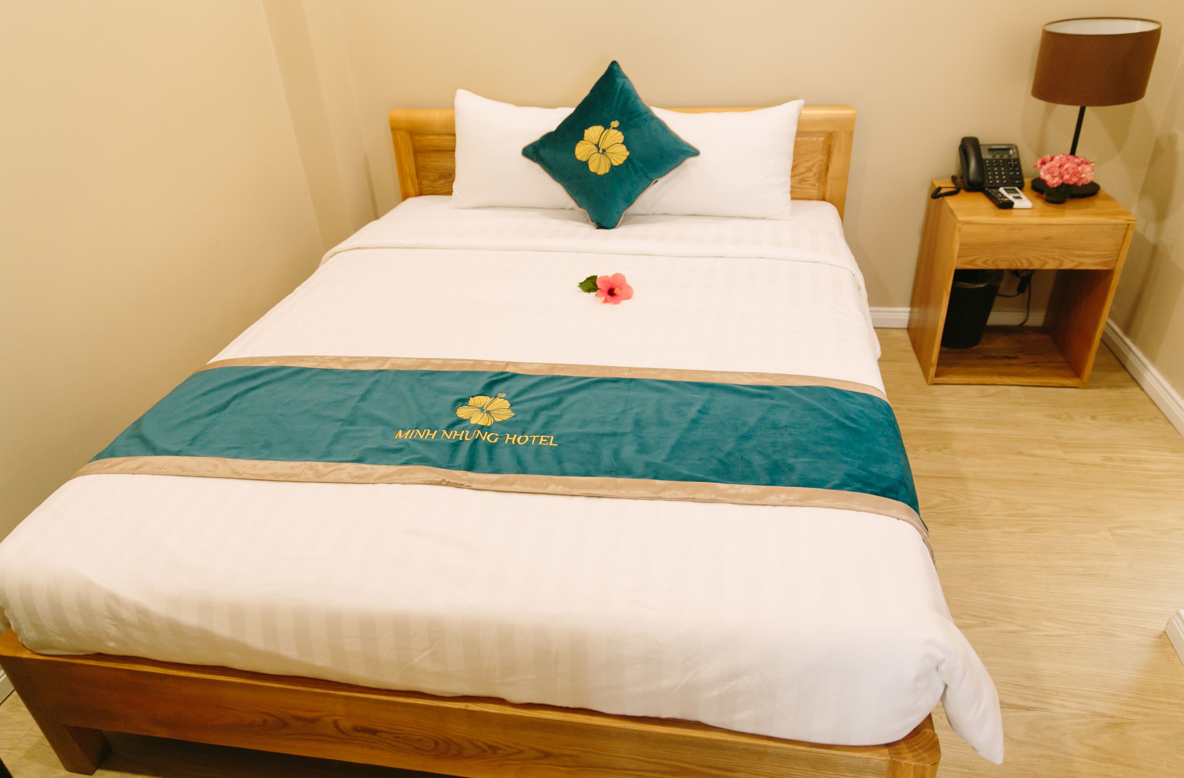 Un lit bien fait, recouvert d’une couvre-lit turquoise et d’une couette blanche, une table de chevet en bois avec une lampe, et une petite fleur posée sur le lit.