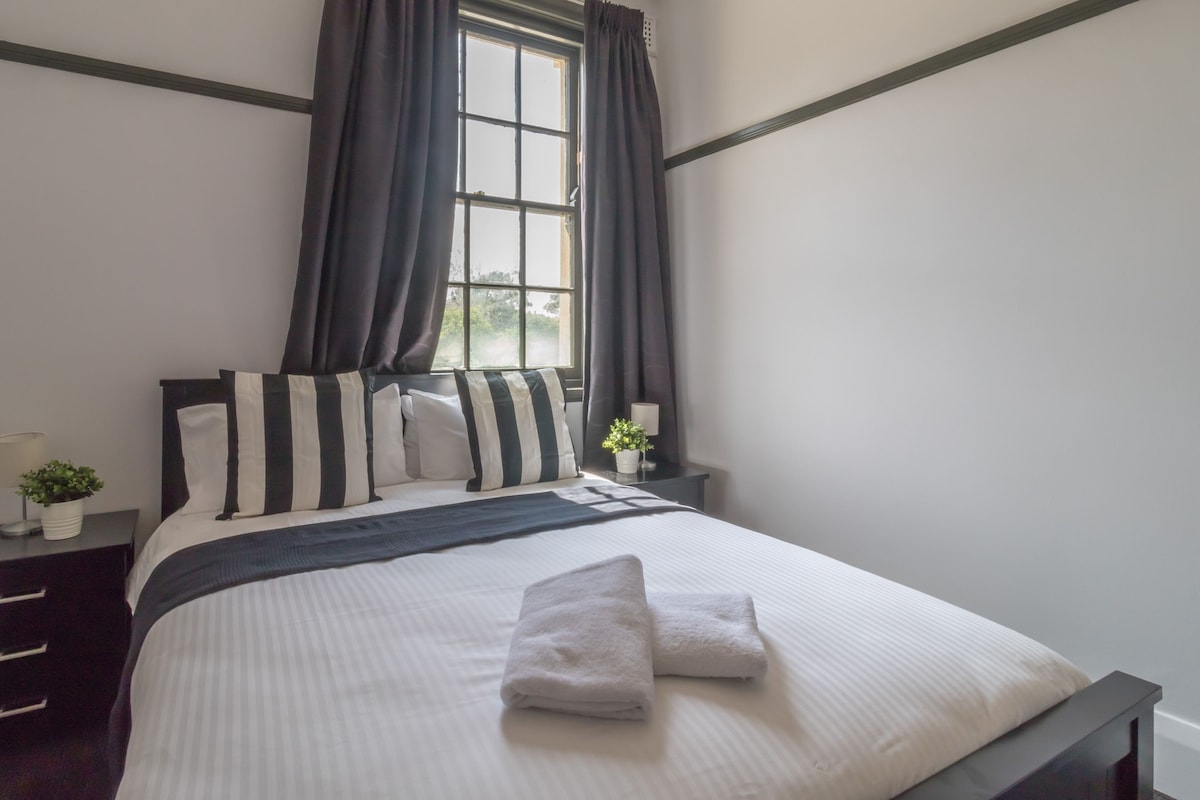Una cama bien hecha con almohadas a rayas, un edredón oscuro y toallas dobladas. Hay una ventana con cortinas, una mesita de noche con una lámpara y una pequeña planta.