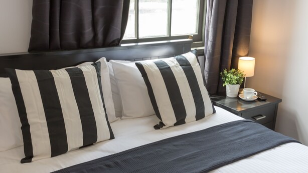 A neatly made bed with striped pillows, a bedside table with a lamp, a potted plant, and a remote control. - Royal Hotel Ryde (Ryde)