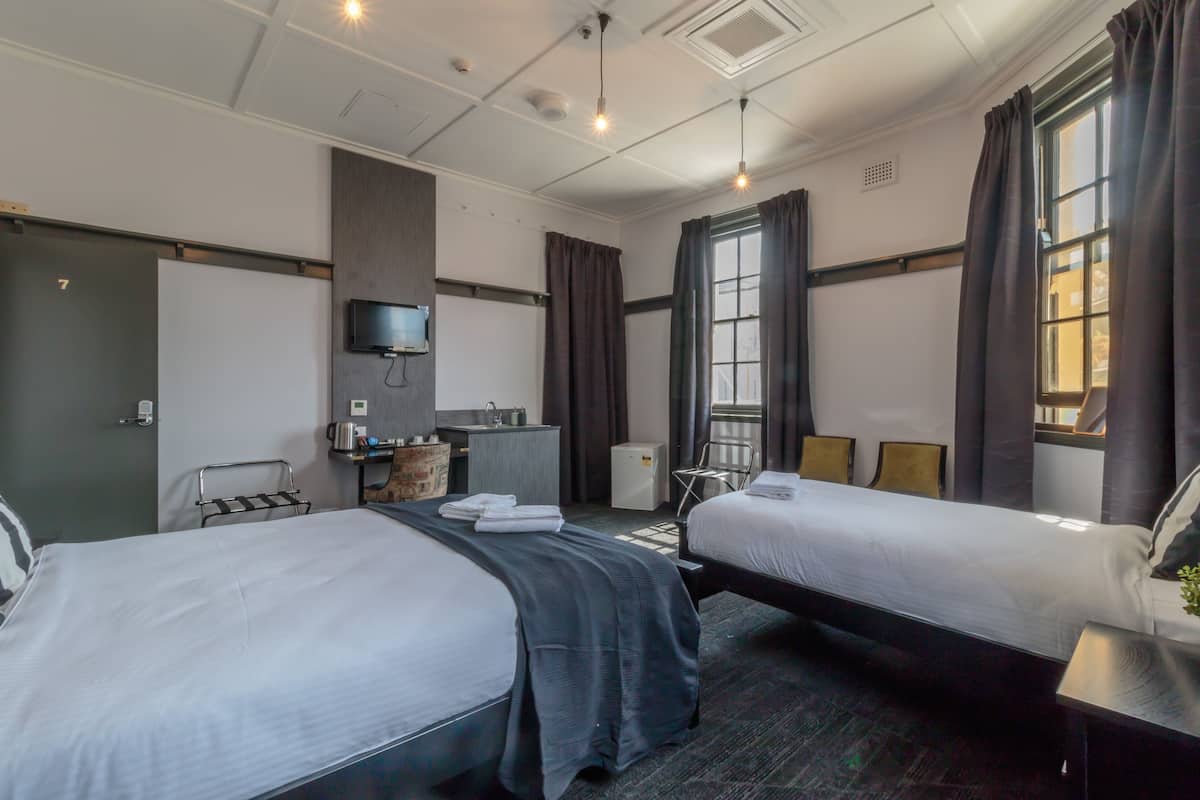 Habitación de hotel con dos camas, un escritorio, una kitchenette pequeña, un televisor montado en la pared y un ventanal con cortinas.