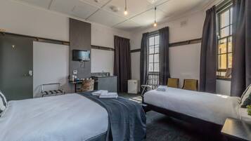 Habitación de hotel con dos camas, un escritorio, una kitchenette pequeña, un televisor montado en la pared y un ventanal con cortinas.